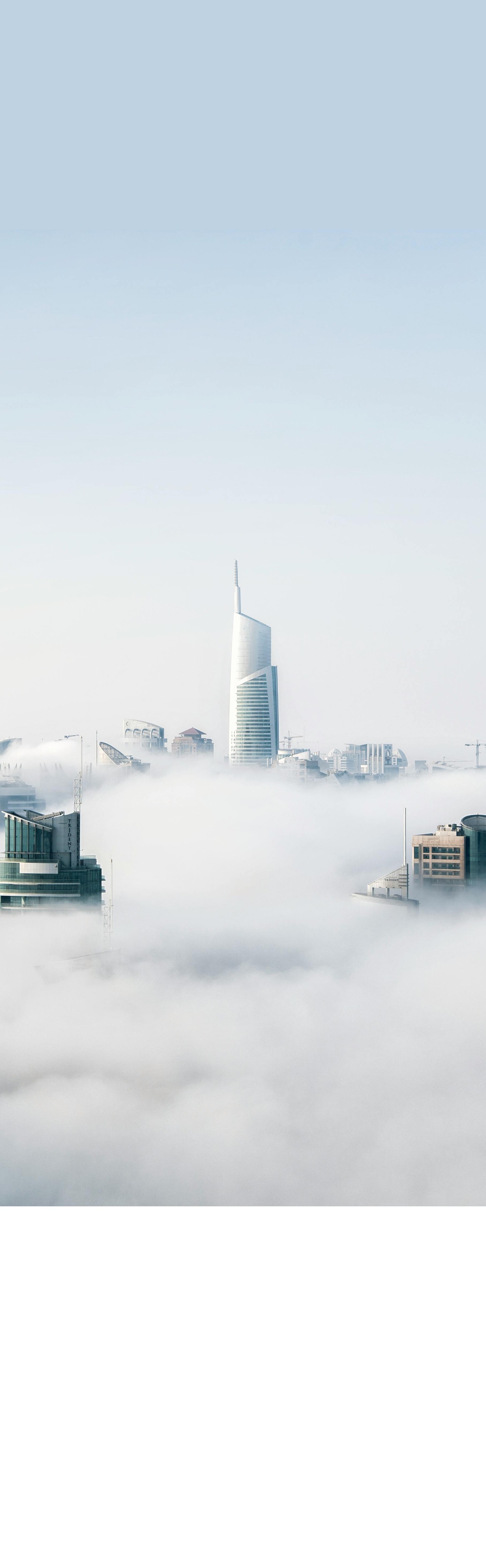 City skyline with skyscrapers emerging through thick fog under a pale blue sky.