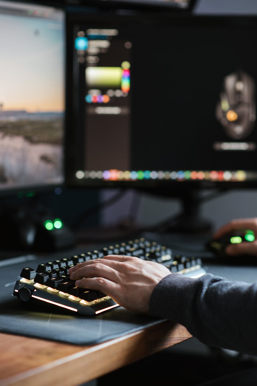 Close-up of a person’s hands using a mechanical keyboard and mouse with colorful backlighting in front of dual computer monitors.