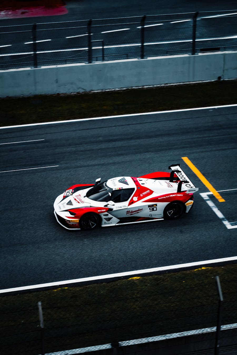 Red and white race car number 90 driving on a racetrack near a concrete barrier and safety fence.